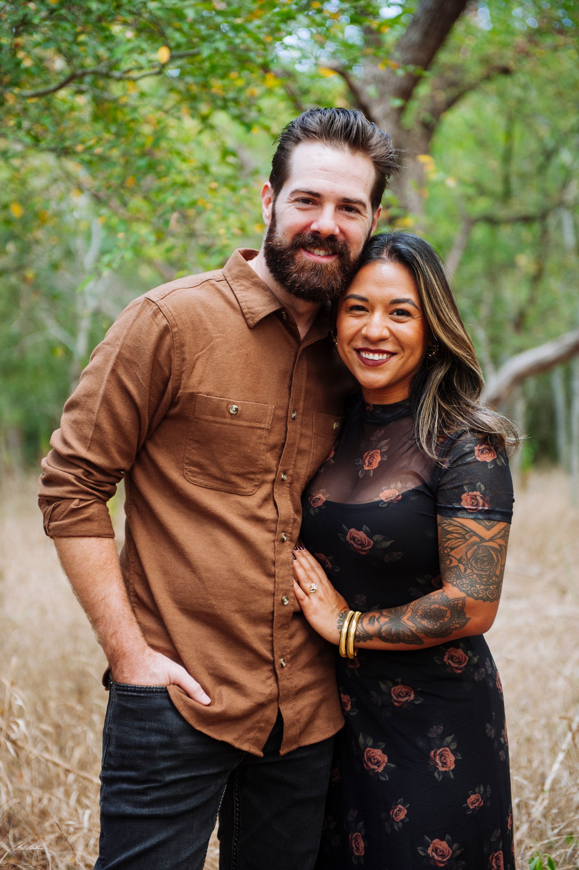 A smiling couple stands close together outdoors amid green trees.