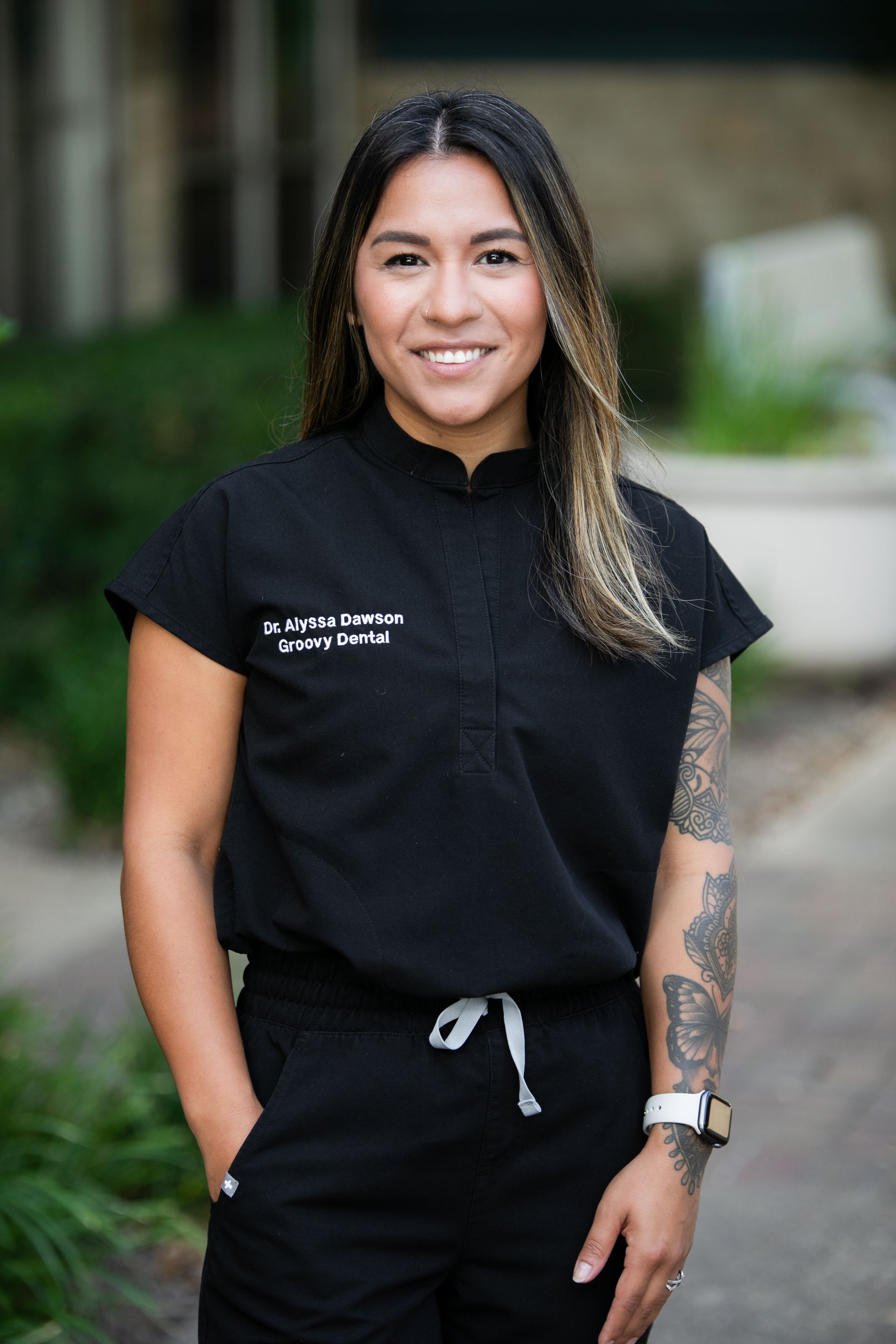Woman standing outdoors in black shirt and pants, smiling with hands in pockets.
