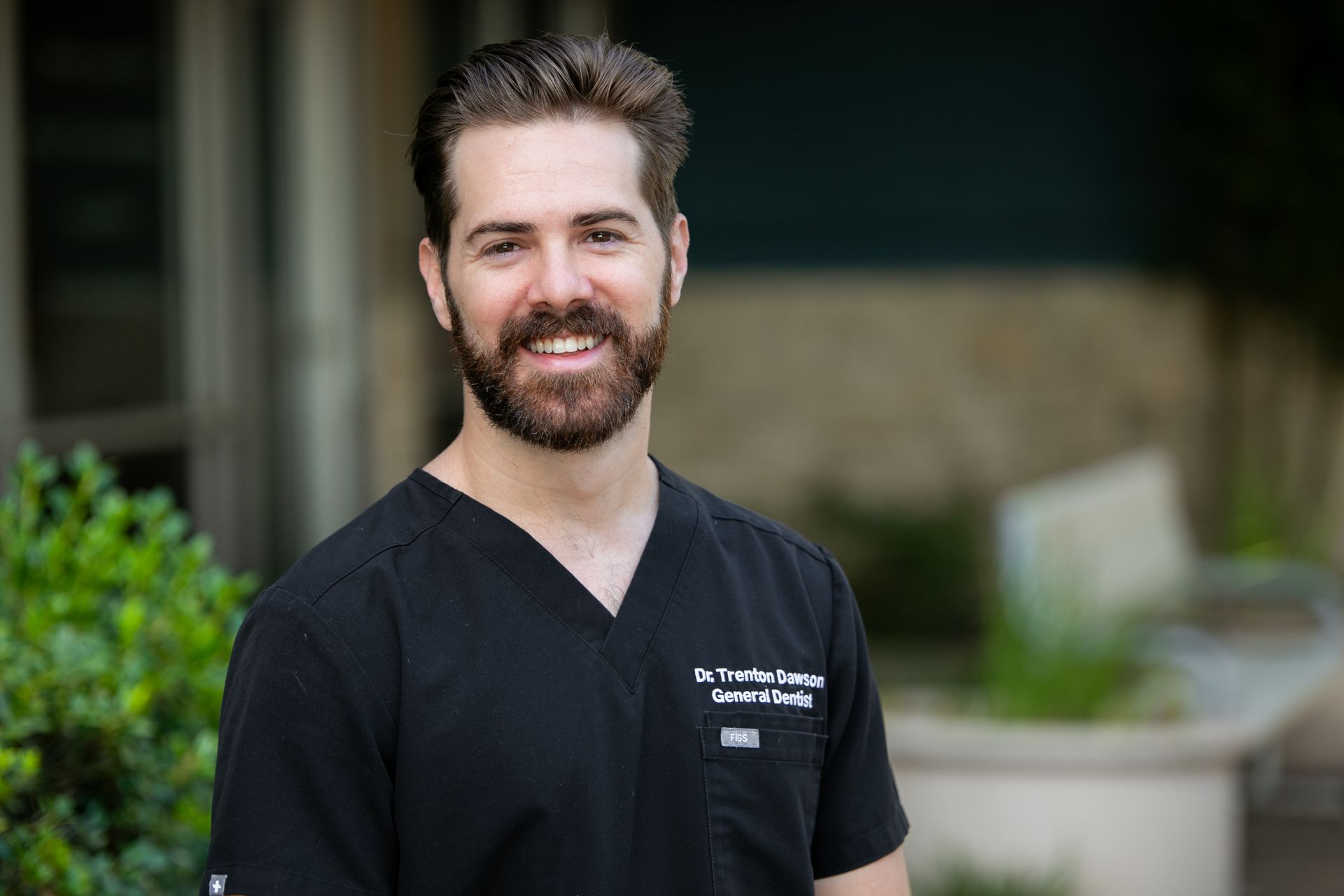 Smiling person in black medical scrubs standing outdoors with greenery in the background
