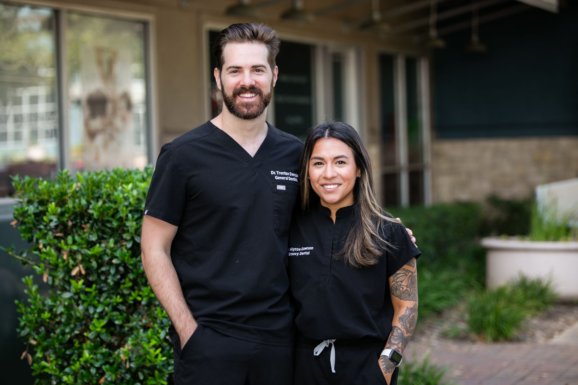 Two smiling coworkers in black scrubs standing outside a building.