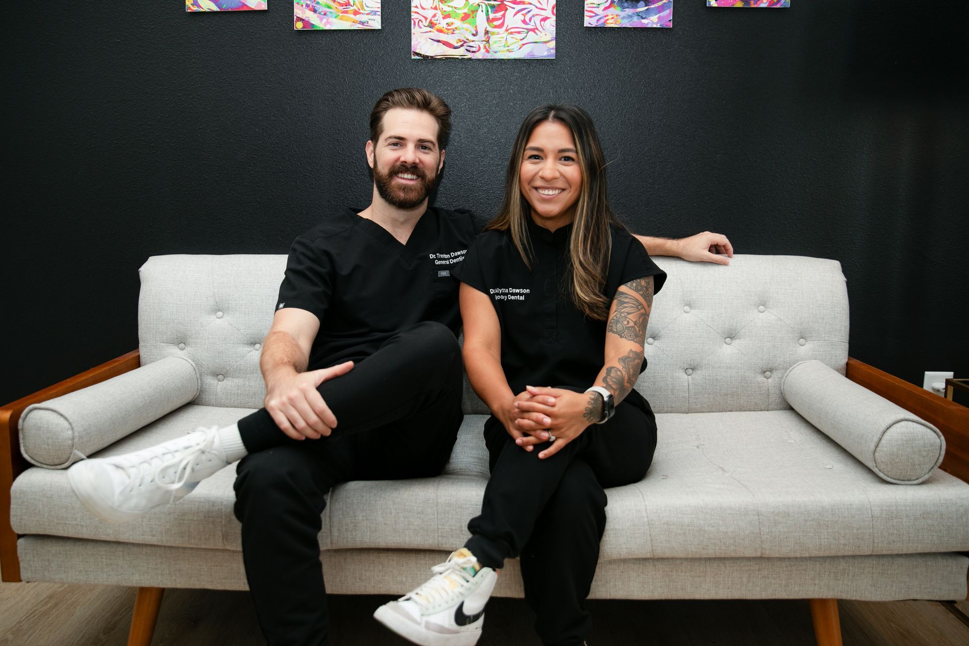 Two people seated on a light gray couch in a black-walled room, smiling at the camera.
