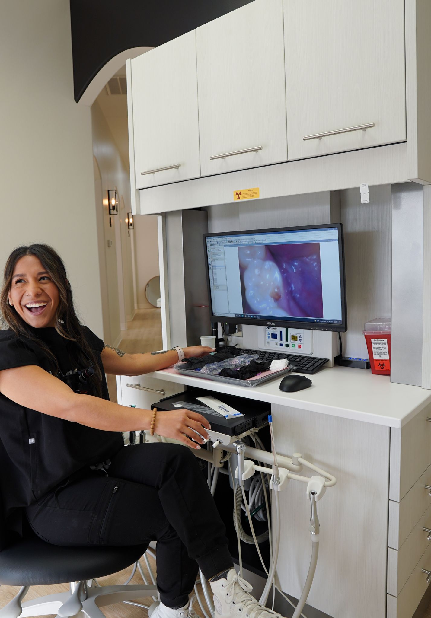 Smiling person at a desktop workstation with a monitor, keyboard, and medical equipment in a clinic cubicle