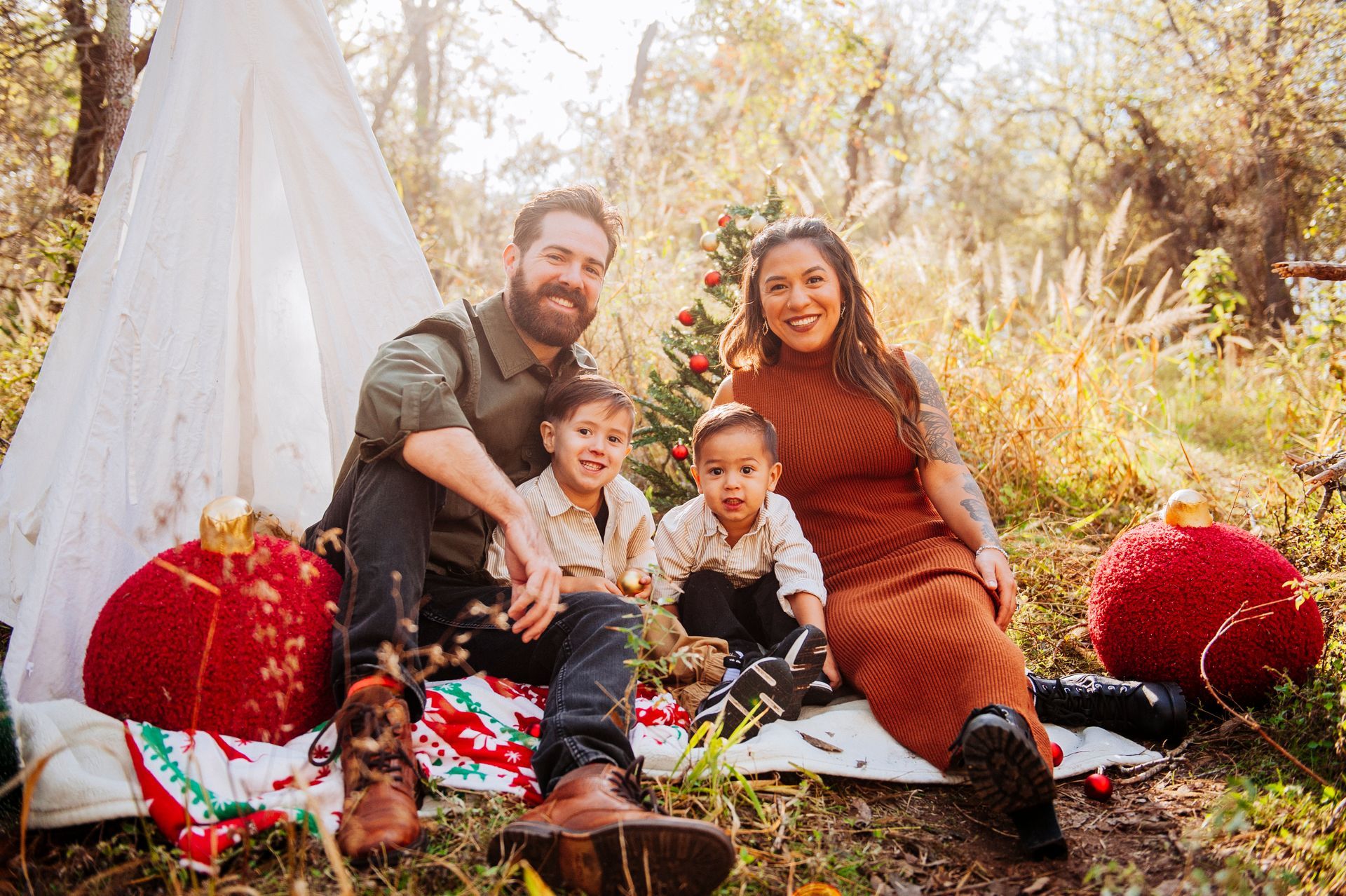 Family sitting on a blanket in a sunlit outdoor picnic scene with a white teepee and red decorations