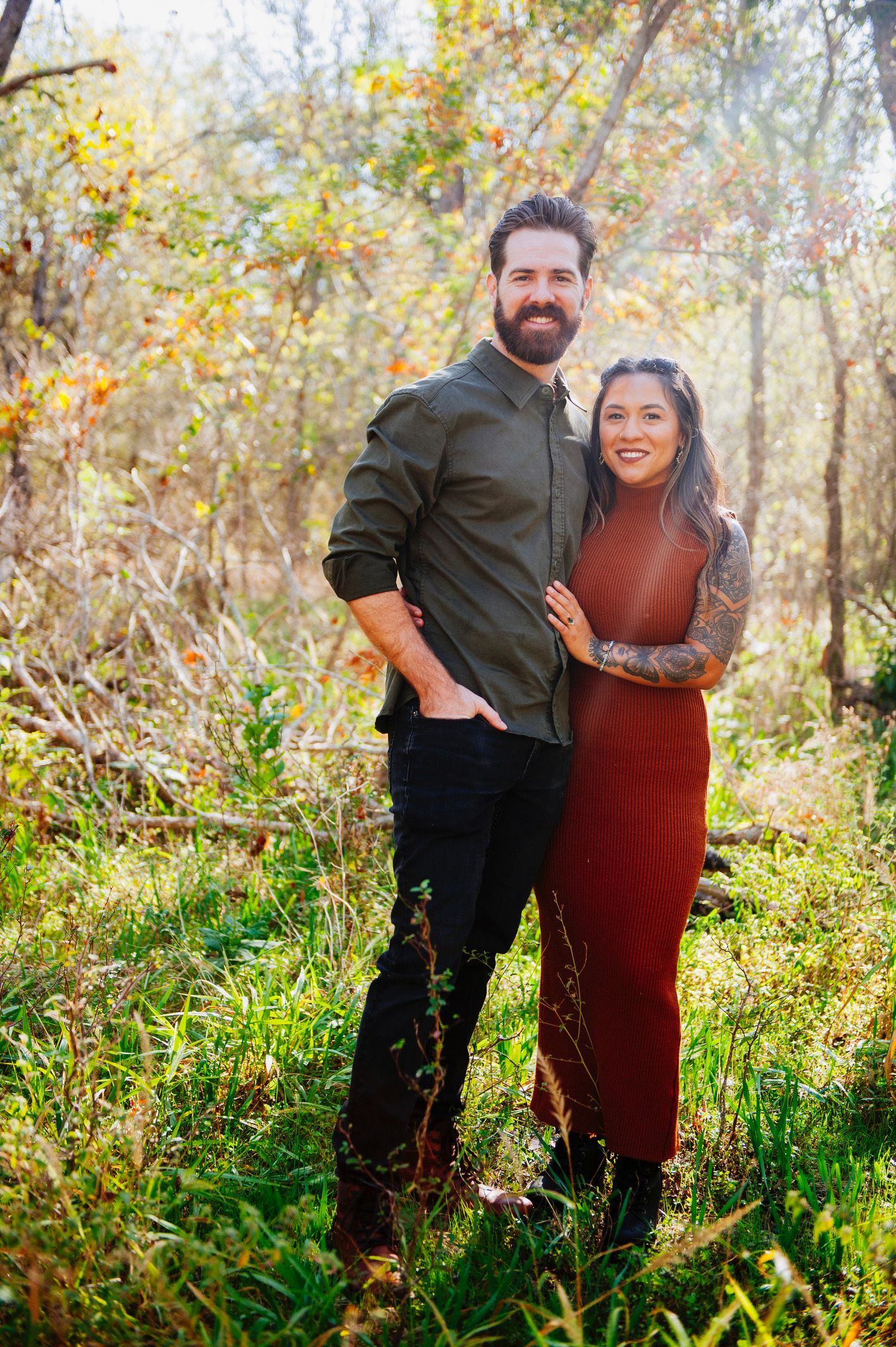 Couple posing outdoors in a sunlit field, with autumn trees and greenery behind them.
