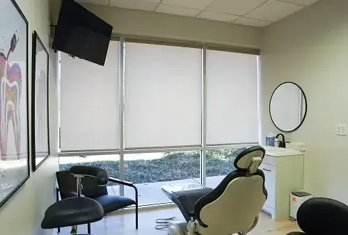 Dental exam room with black chairs, wall monitor, round mirror, and large window blinds