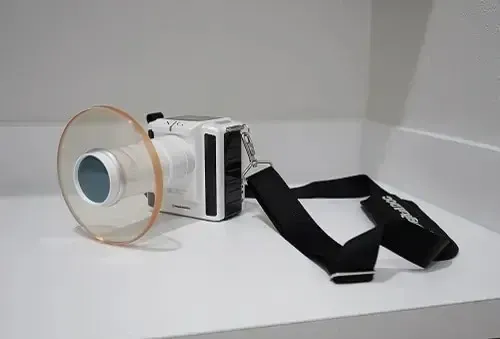 White camera with a large lens attached to a black shoulder strap on a plain white surface.