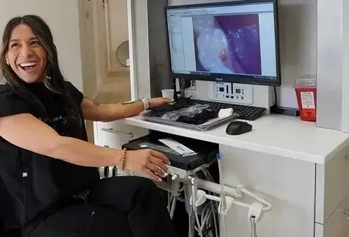 Woman smiling at a workstation with a large monitor showing a medical scan and equipment beneath the desk