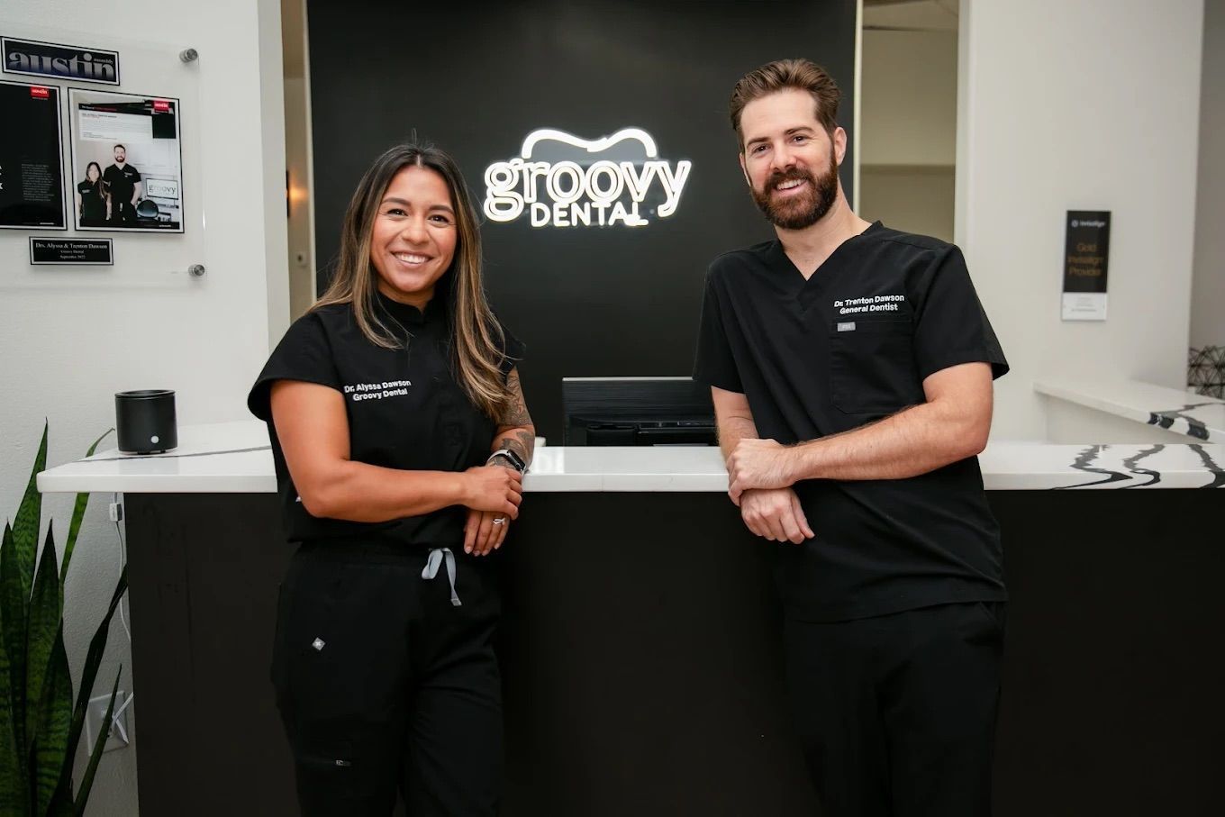 Two smiling staff in black uniforms stand behind a counter with a Groovy Den sign.