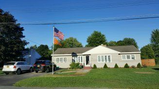 Suburban house with American flag, cars in driveway, and green lawn under a blue sky.