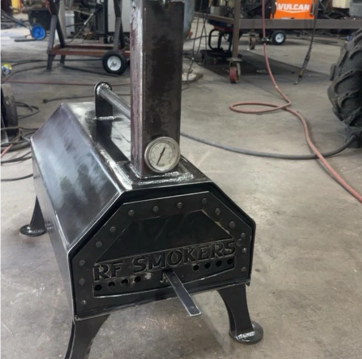 Black metal RF smoker with a smokestack, temperature gauge, and handle in a workshop.
