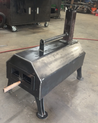 Welded steel pizza oven with flue, door, handle, and legs, sitting on concrete floor.