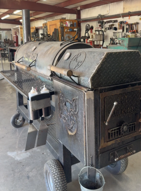 A large, black, custom-built smoker with a grill, used in a workshop setting.