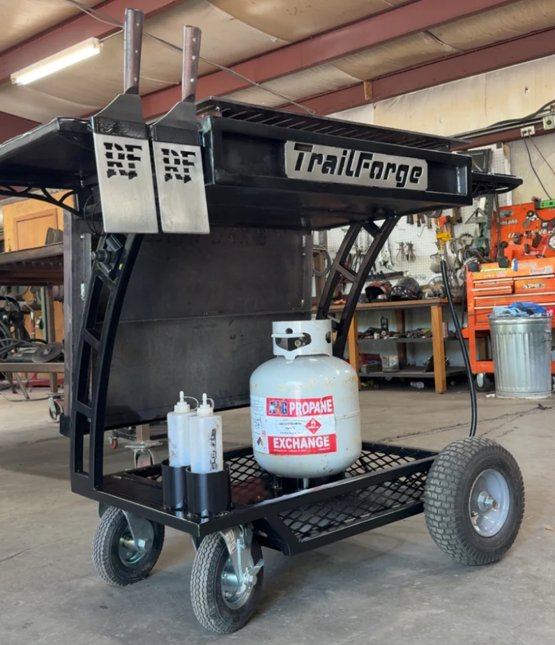 Black metal TrailForge grilling cart with propane tank, spatulas, and oil bottles.