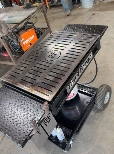 Black Trailgator BBQ grill cart with diamond plate surfaces, a propane tank, and a metal grated grill.