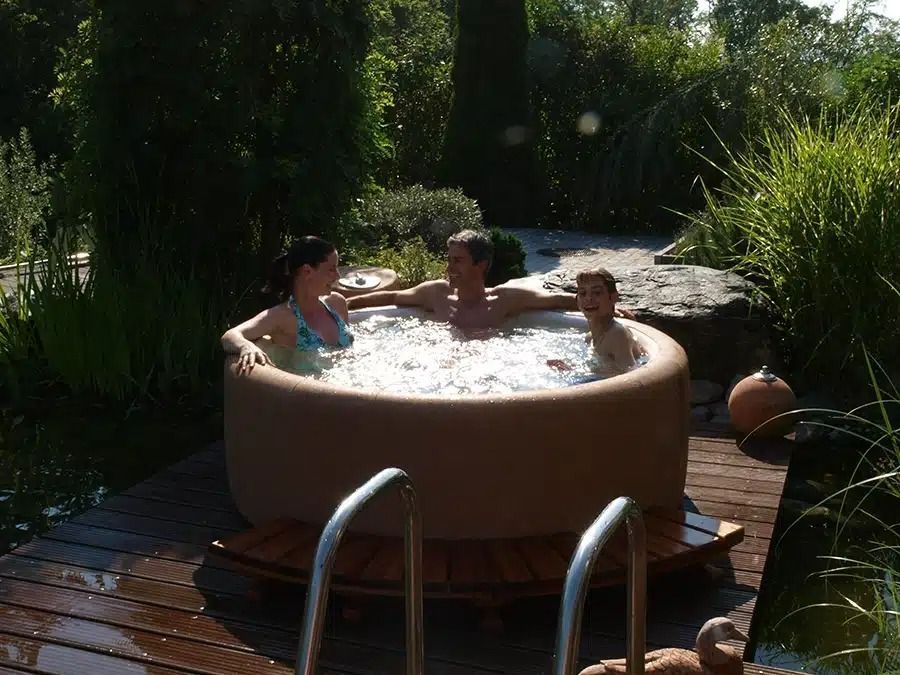 Three people relaxing in a round outdoor hot tub surrounded by lush greenery.