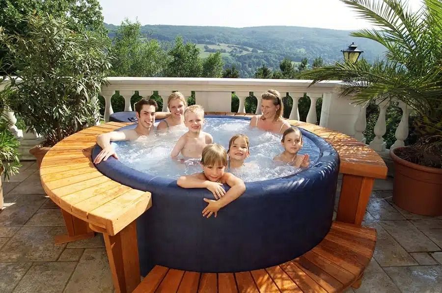 Family of seven in a round hot tub on a terrace, overlooking a scenic landscape.