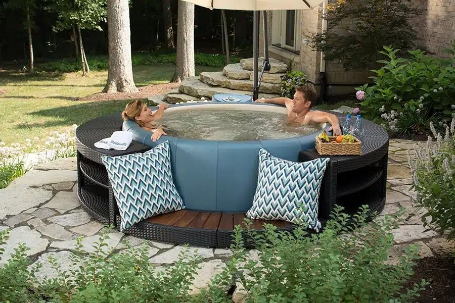 Couple relaxing in a round outdoor hot tub with teal pillows, drinks, and an umbrella.