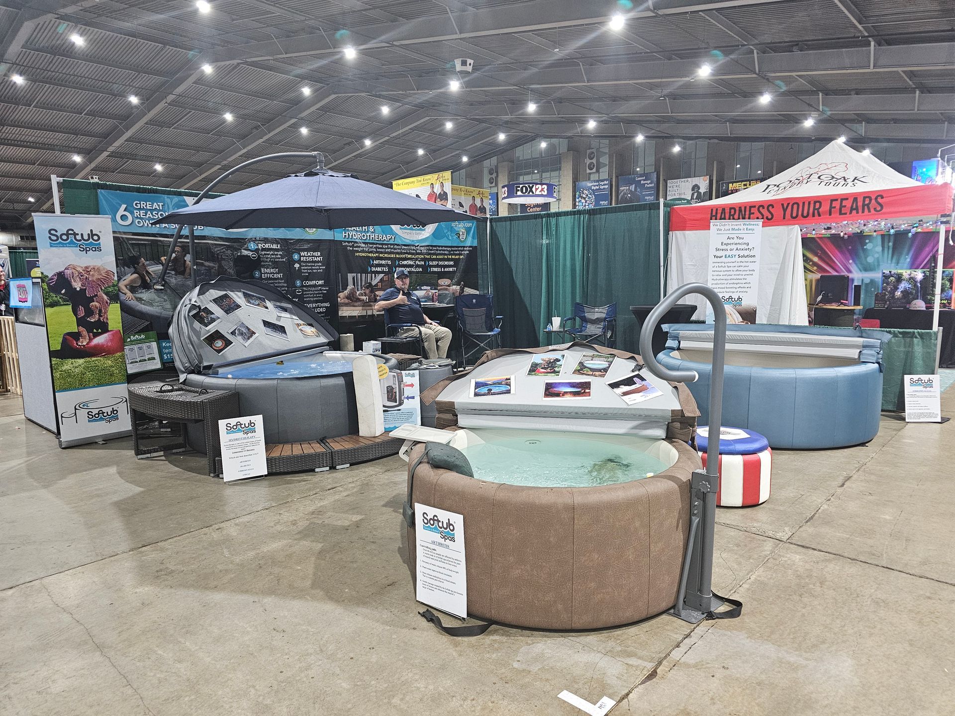 Indoor trade show booth showcasing hot tubs. Booths display various models, a large umbrella, and signage.