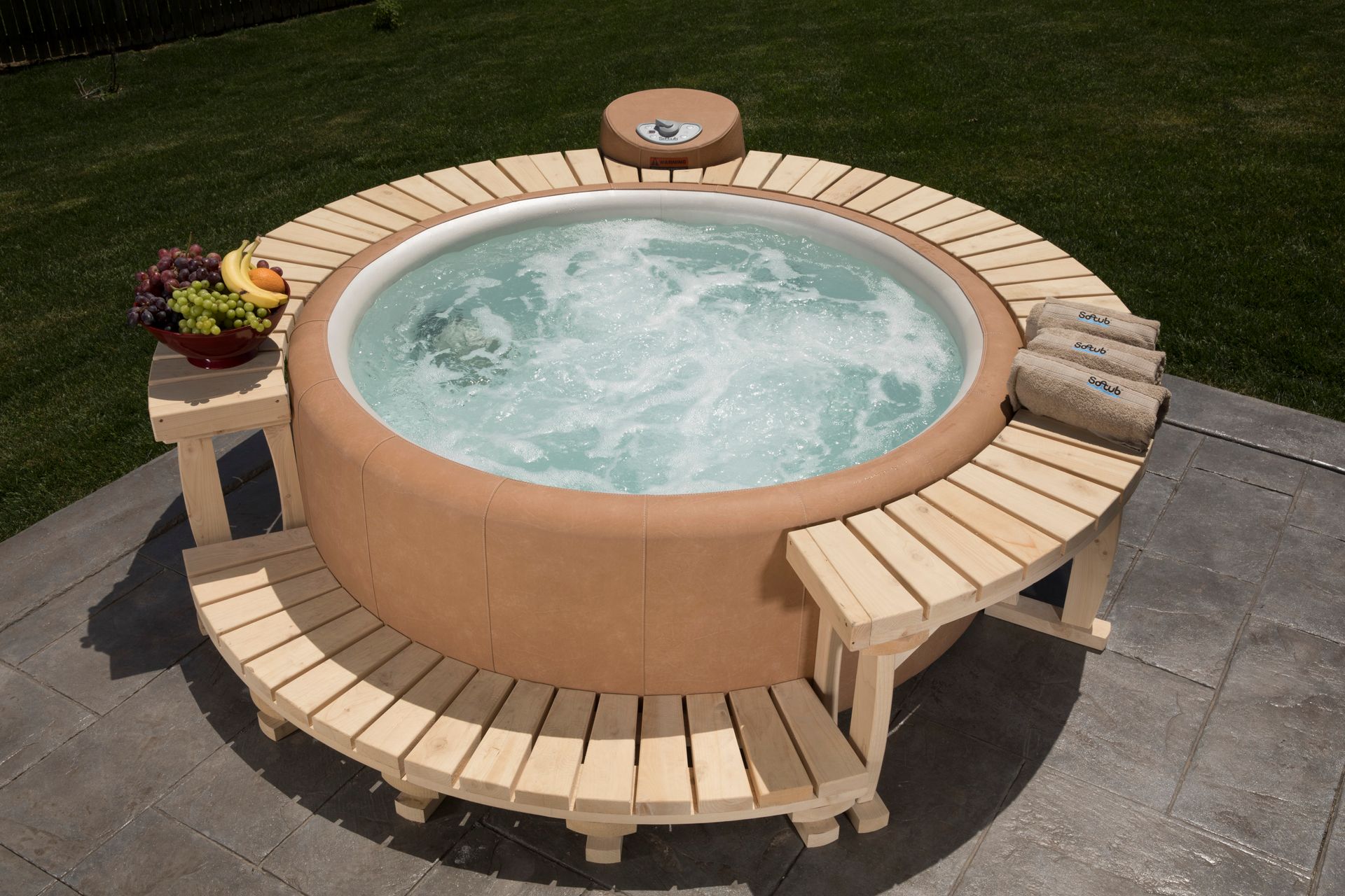 Round hot tub with wooden surround, set outdoors on a patio with grass, fruit, and towels.