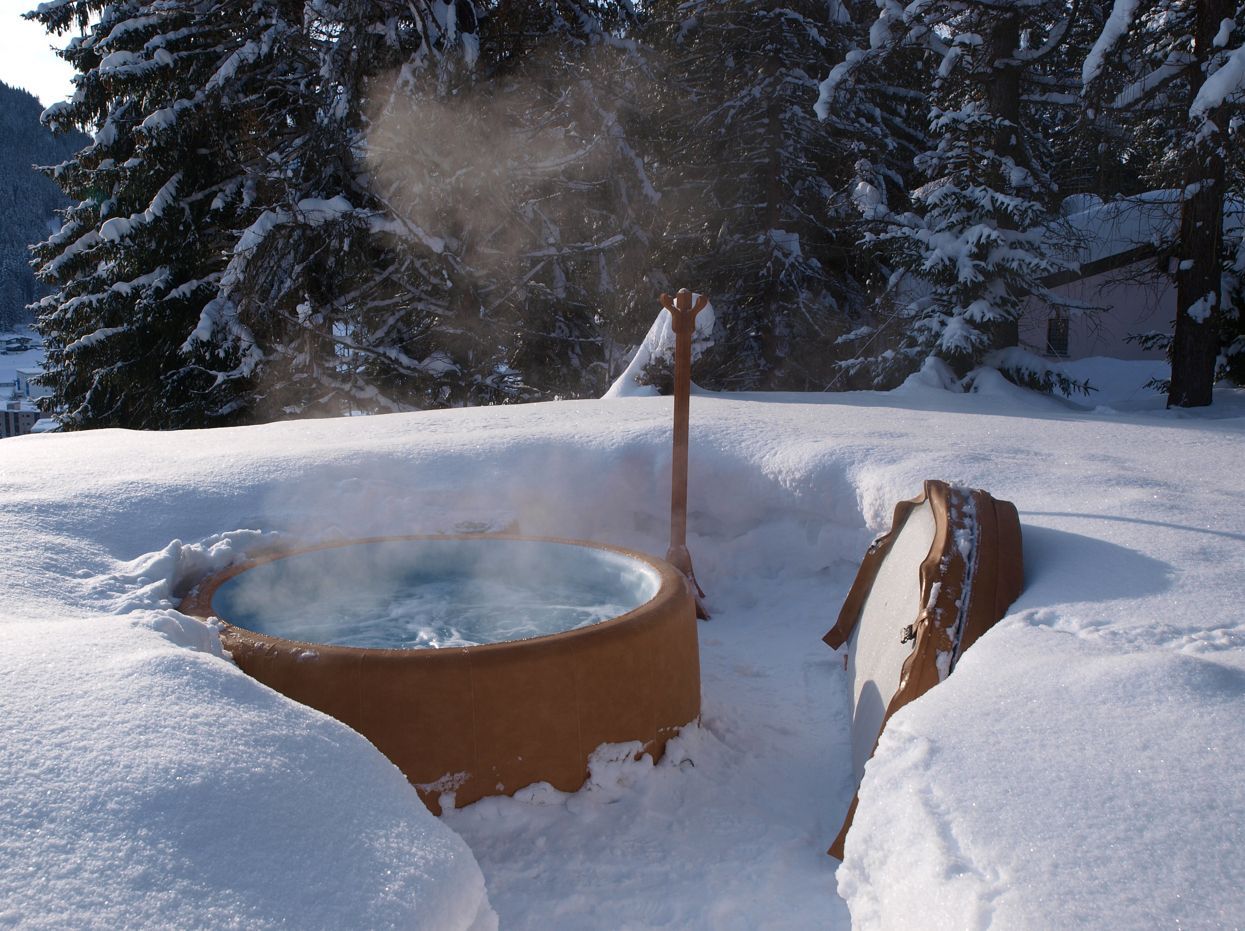 Hot tub steaming in the snow, surrounded by snow banks. Mountains and trees in the background.