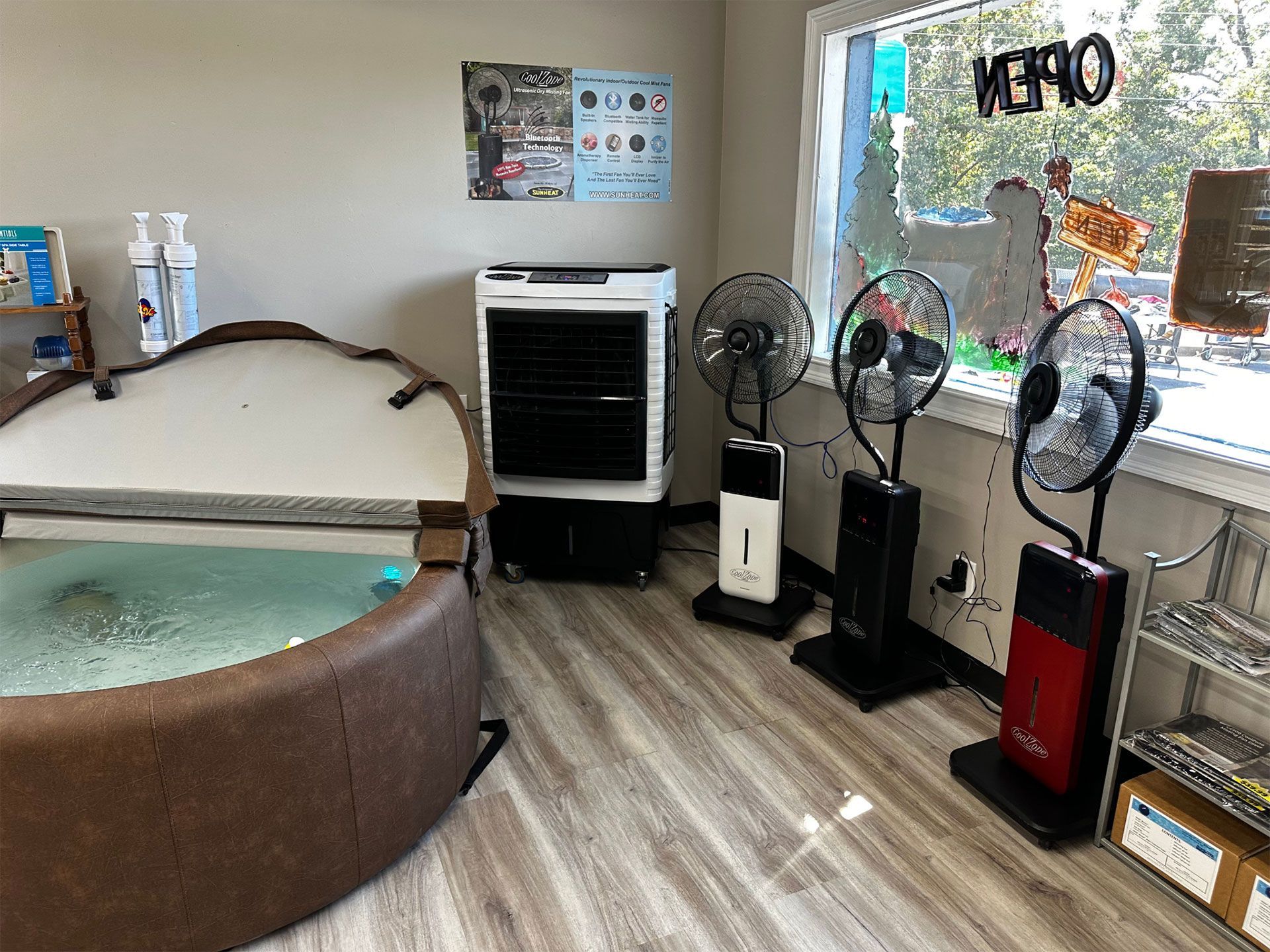 A hot tub, cooling unit, and fans sit near a window in a shop with wooden flooring.