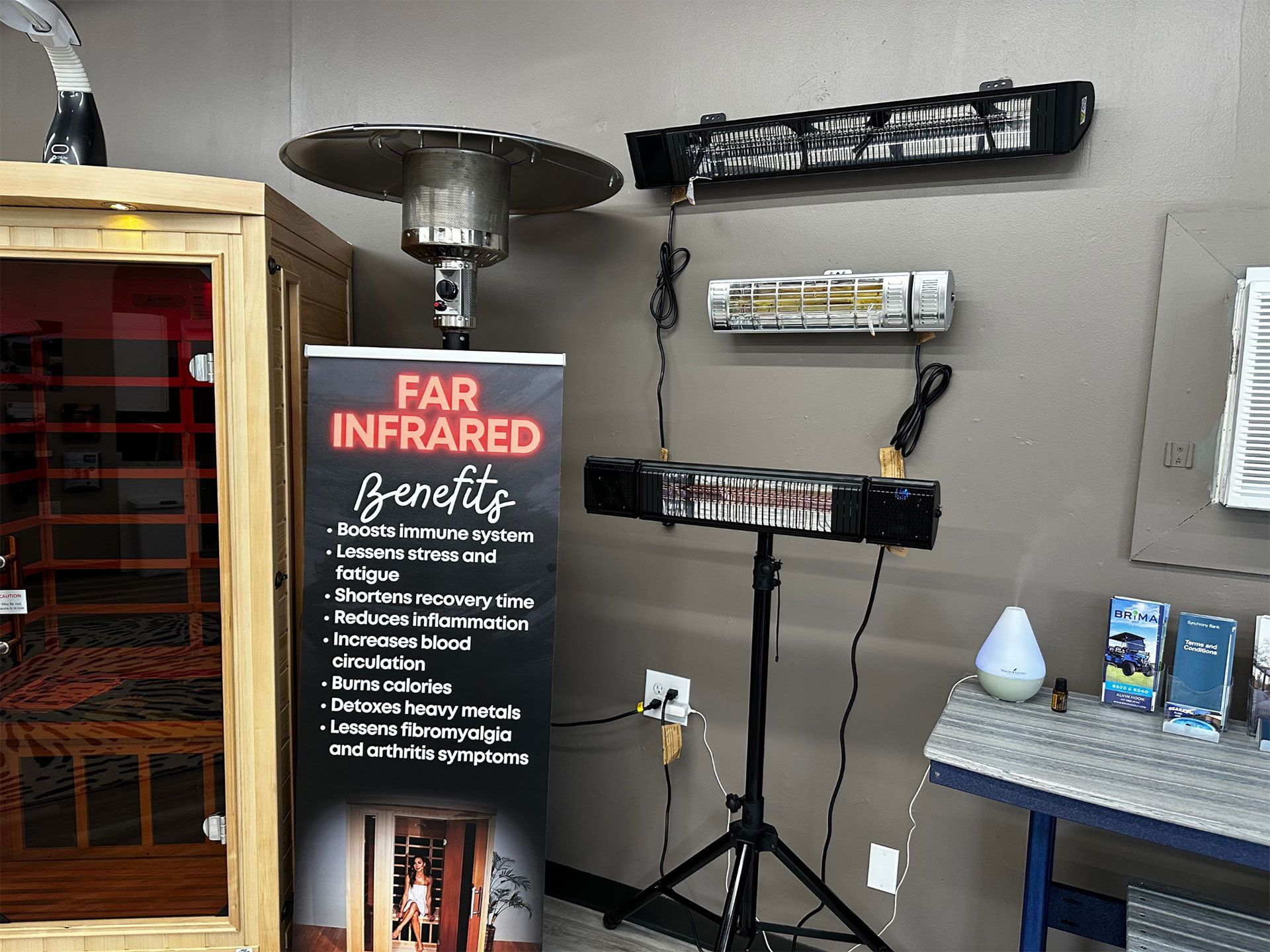 Display of infrared heaters and a sauna. A banner lists benefits; a diffuser and products are on a table.