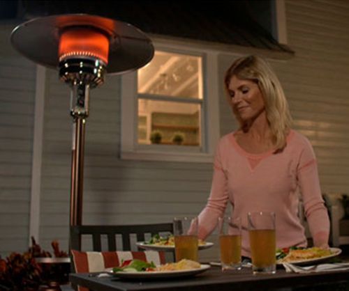 Woman setting a table with food and drinks outdoors at night with a patio heater lit.