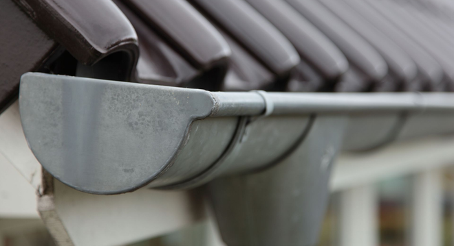 Close-up of a gray rain gutter attached to a dark shingled roofline