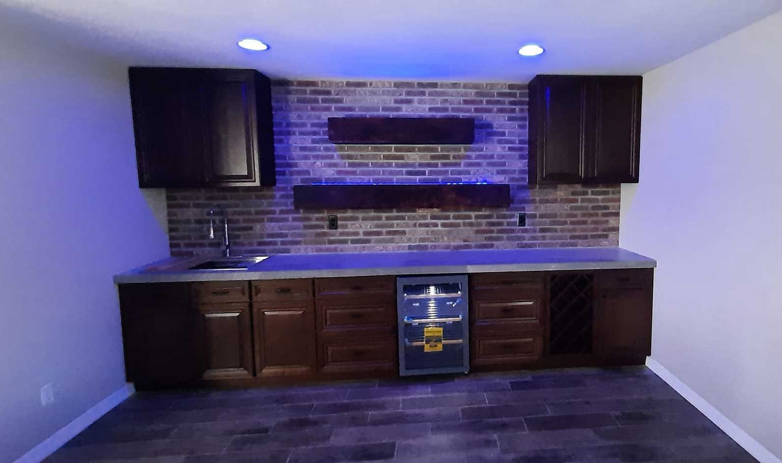 Modern wet bar with dark wood cabinets, brick backsplash, blue lighting, and a central beverage fridge.