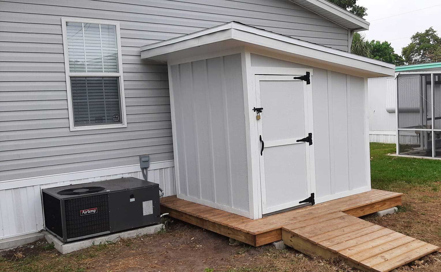 White outdoor shed with double doors and ramp beside a gray house, near a gravel path and AC unit