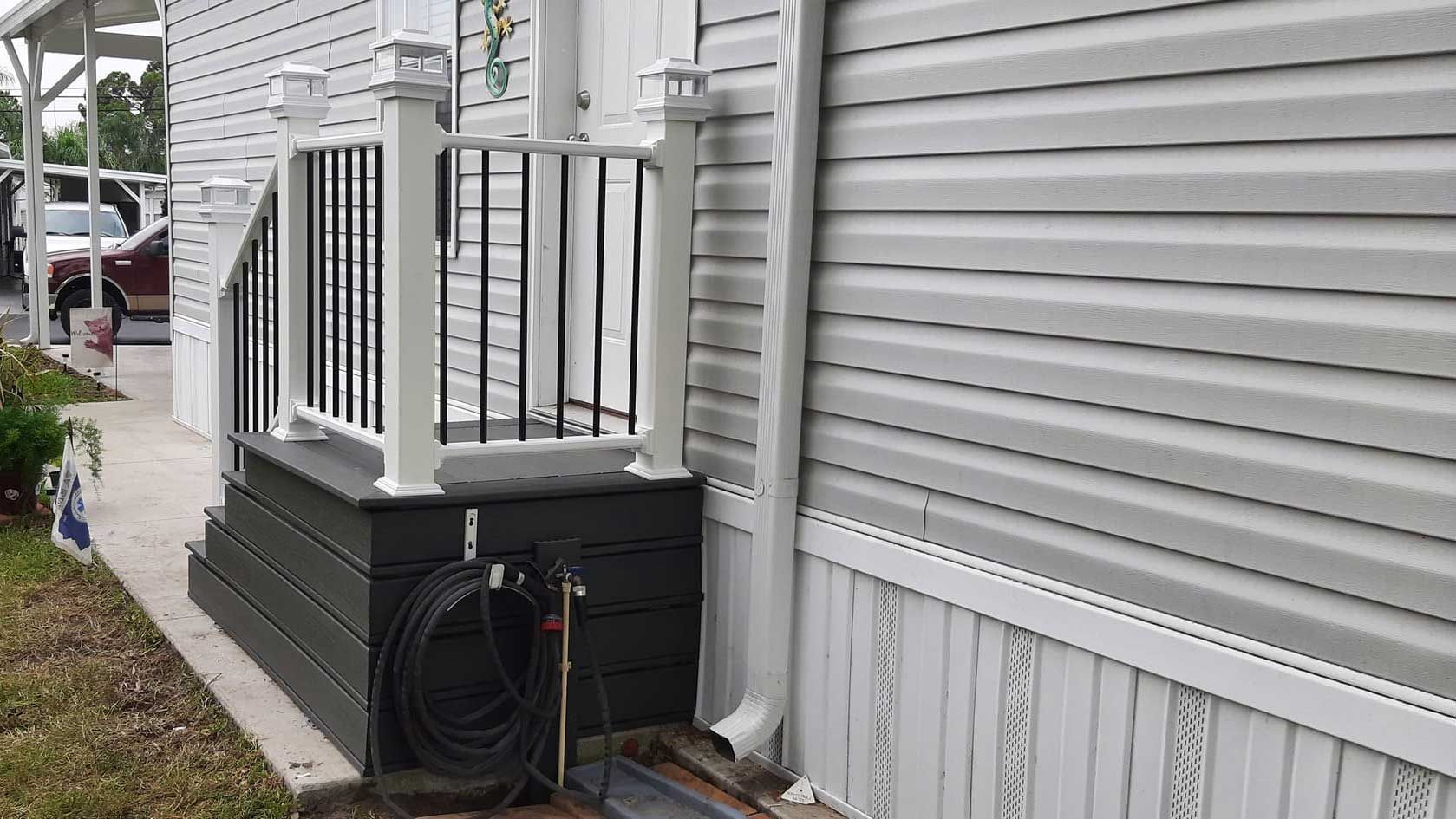 Side of a house with a small porch, white railings, dark siding, and a coiled hose on the ground.