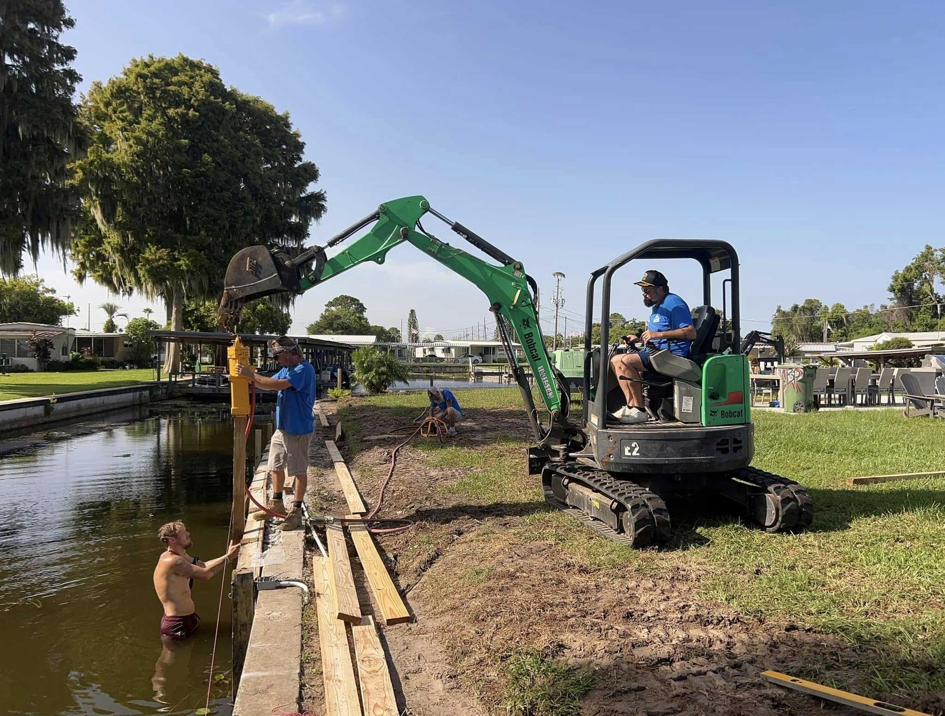 Excavator clears mud beside a pond while workers and a child stand along the canal bank.
