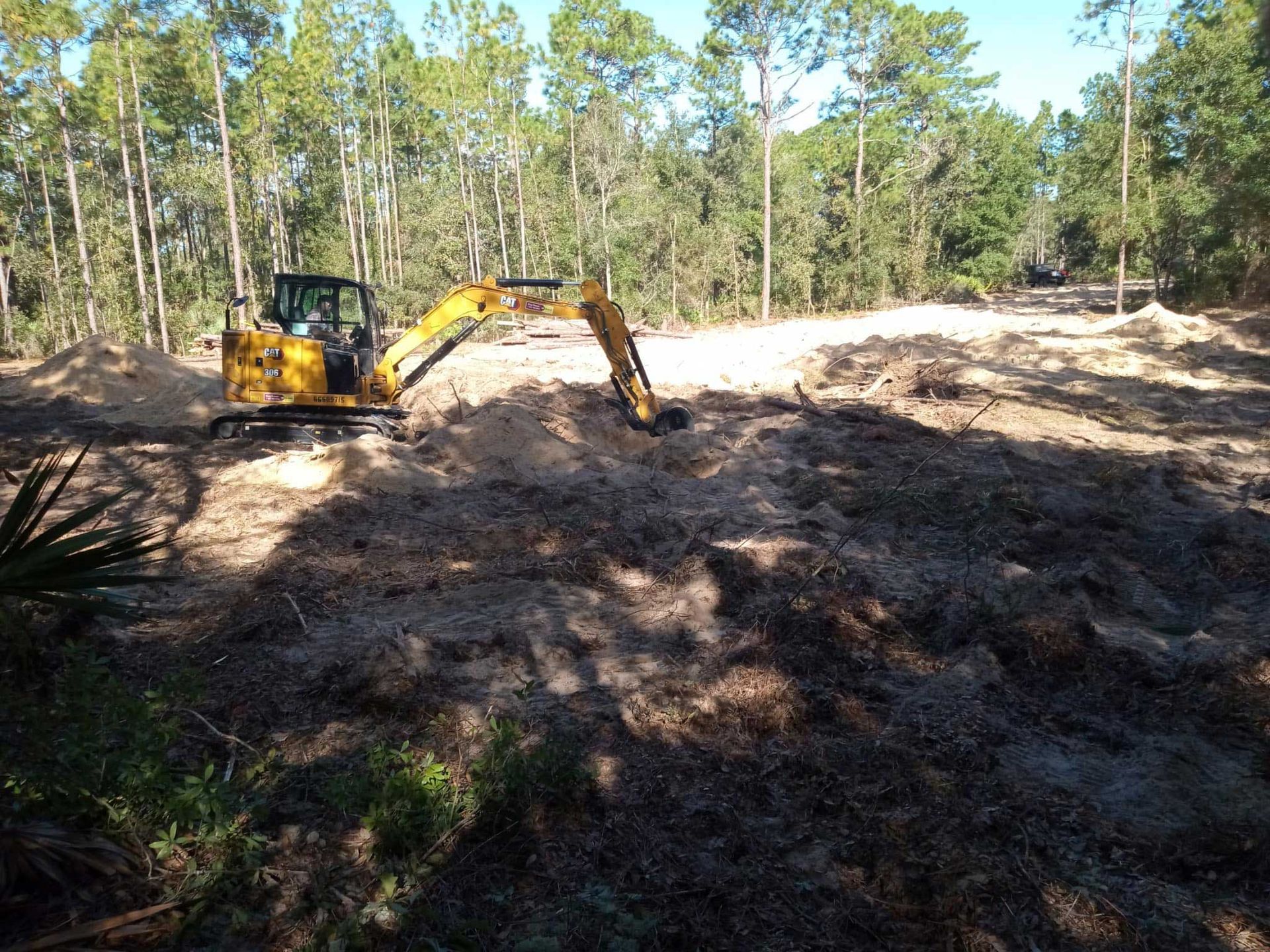 Yellow excavator clearing a dirt lot in a wooded area under bright sunlight