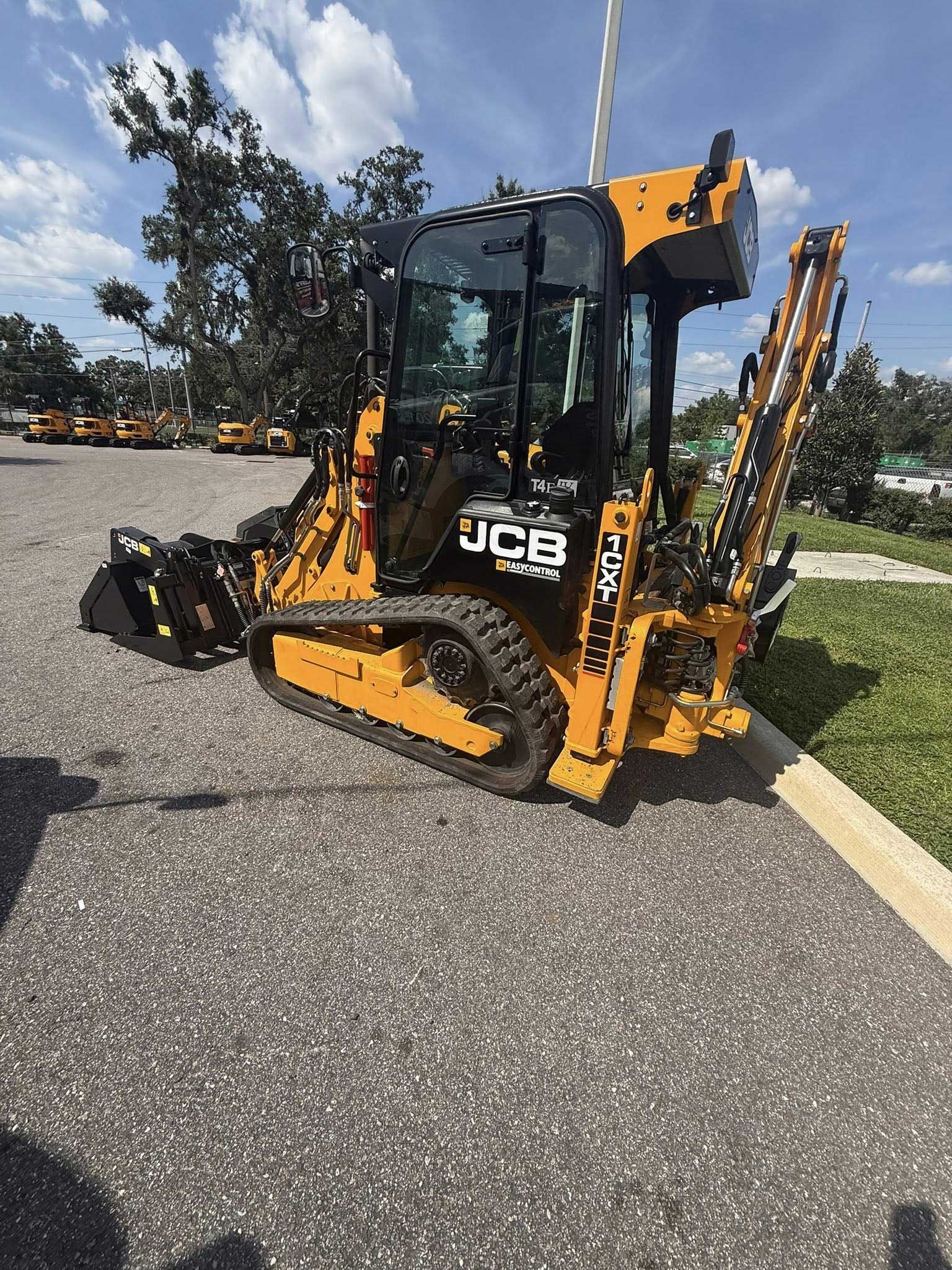 Yellow JCB backhoe loader parked on a gravel lot beside grass and trees.