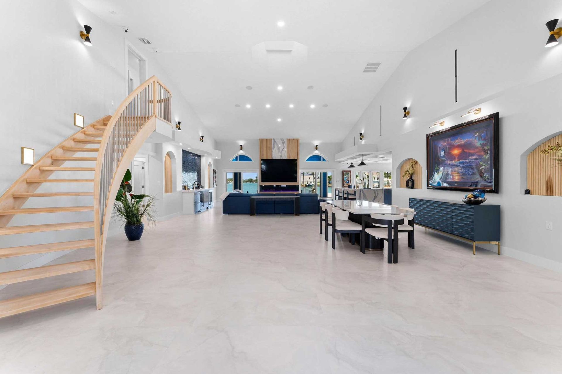 Bright modern living room with curved staircase, white walls, blue sofas, and a large wall-mounted TV
