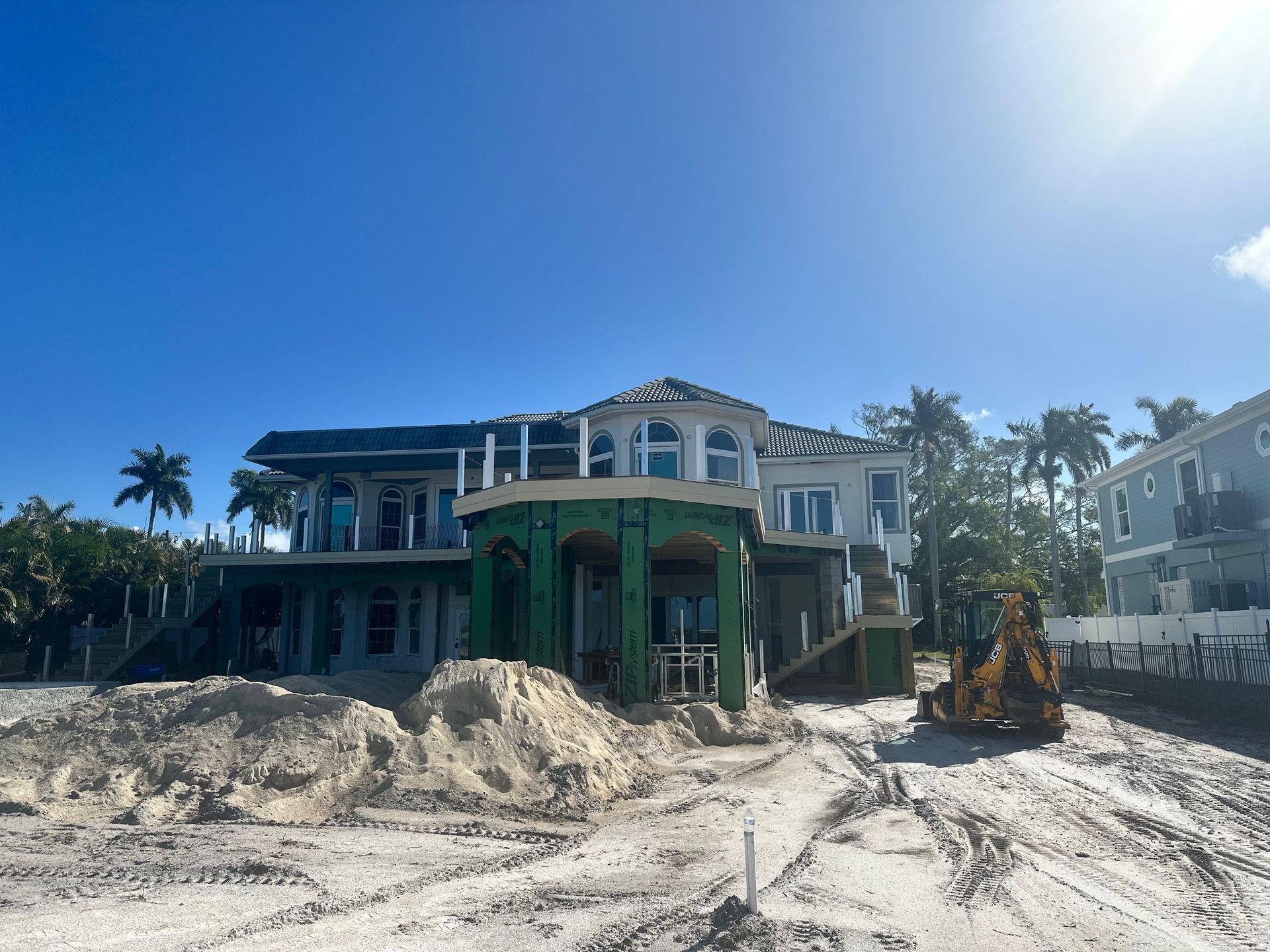 Damaged seaside house under construction with piles of sand and an excavator in front.