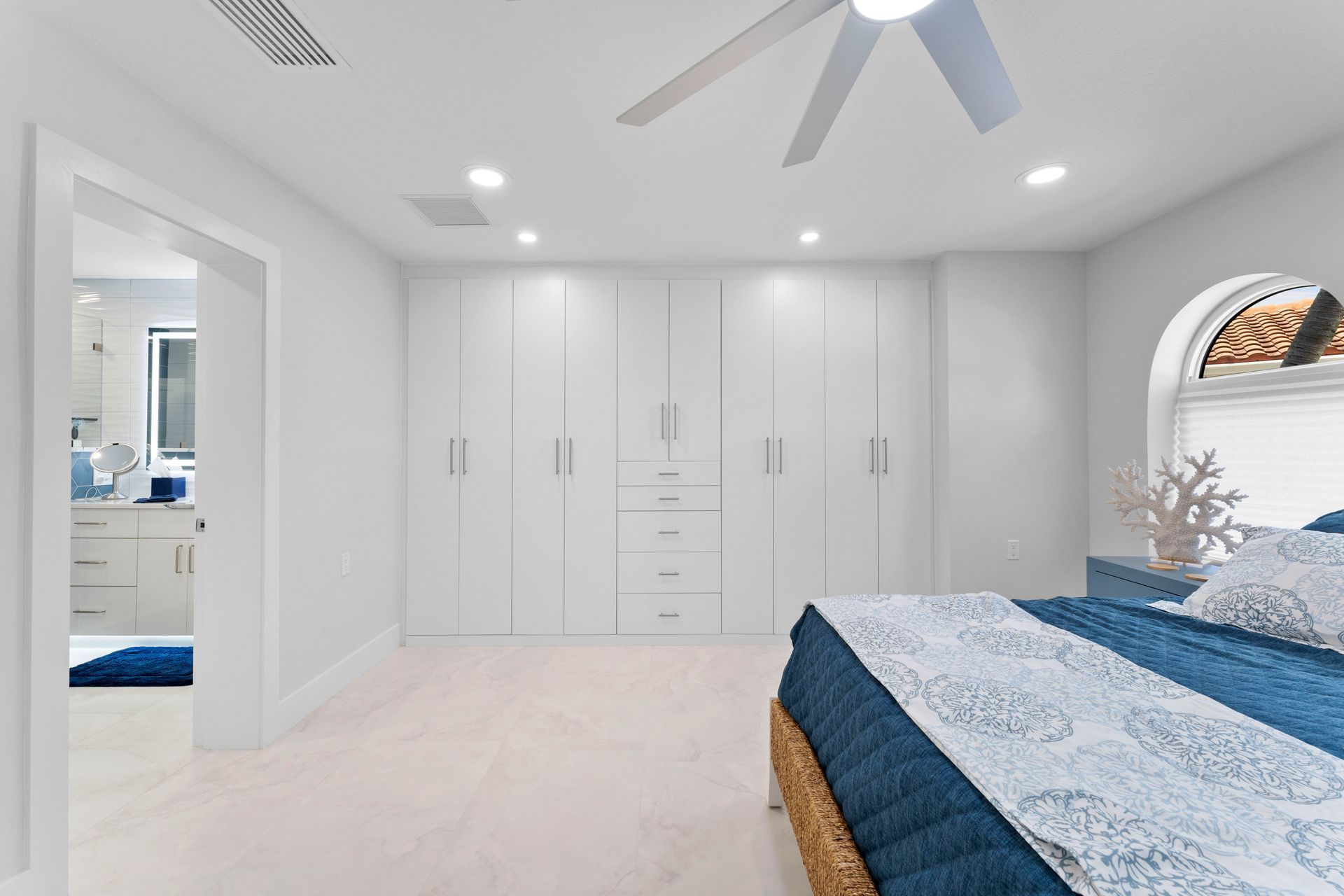 Bright white bedroom with built-in closets, blue bedding, ceiling fan, and an arched window.