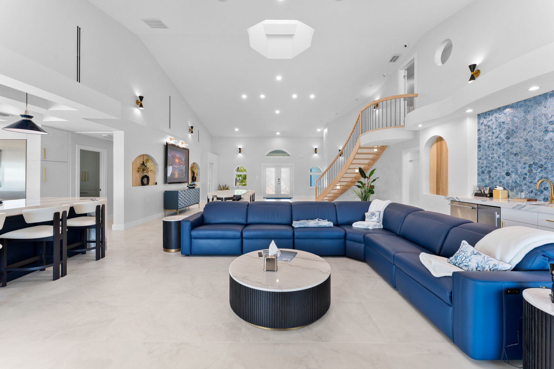 Bright modern open-plan living room with blue sectional sofa, round coffee table, and staircase in the background.