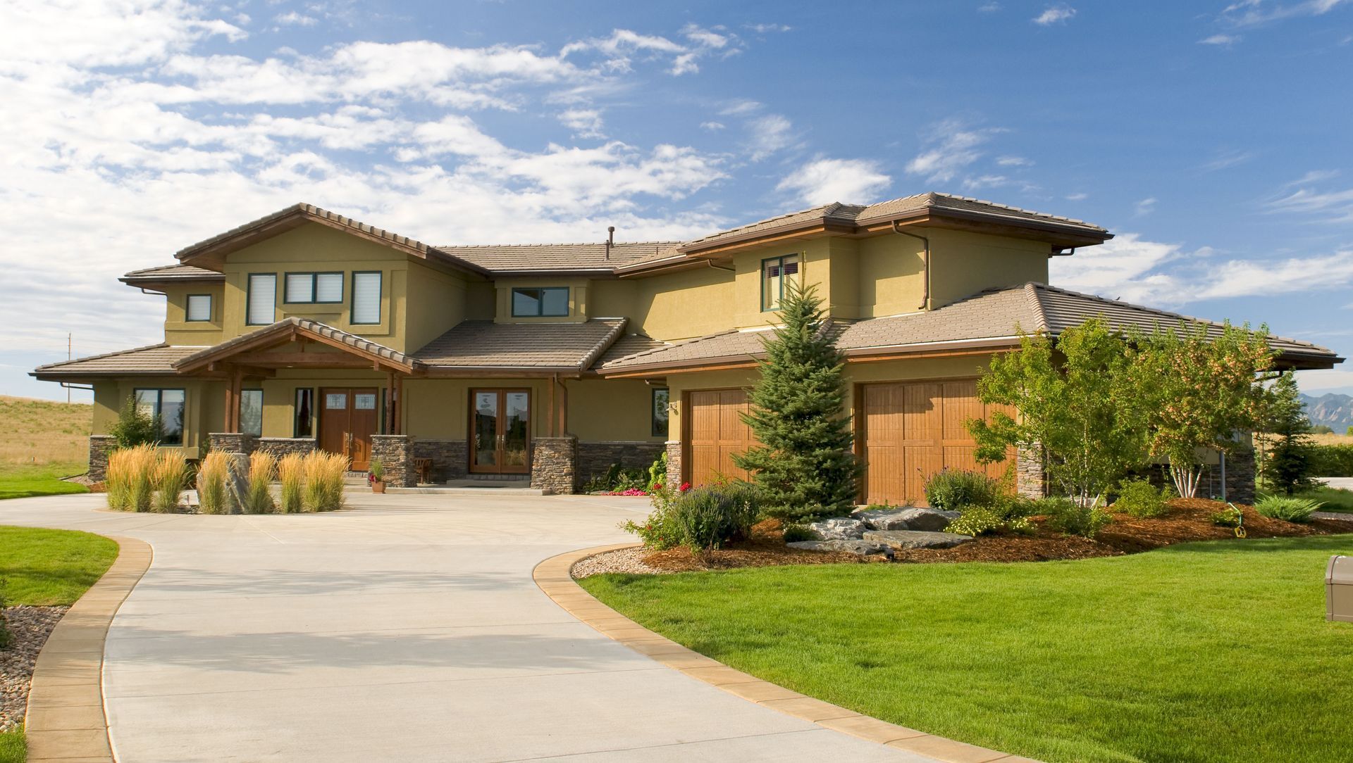 Large modern two-story house with a curved driveway, green lawn, and trees under a partly cloudy sky