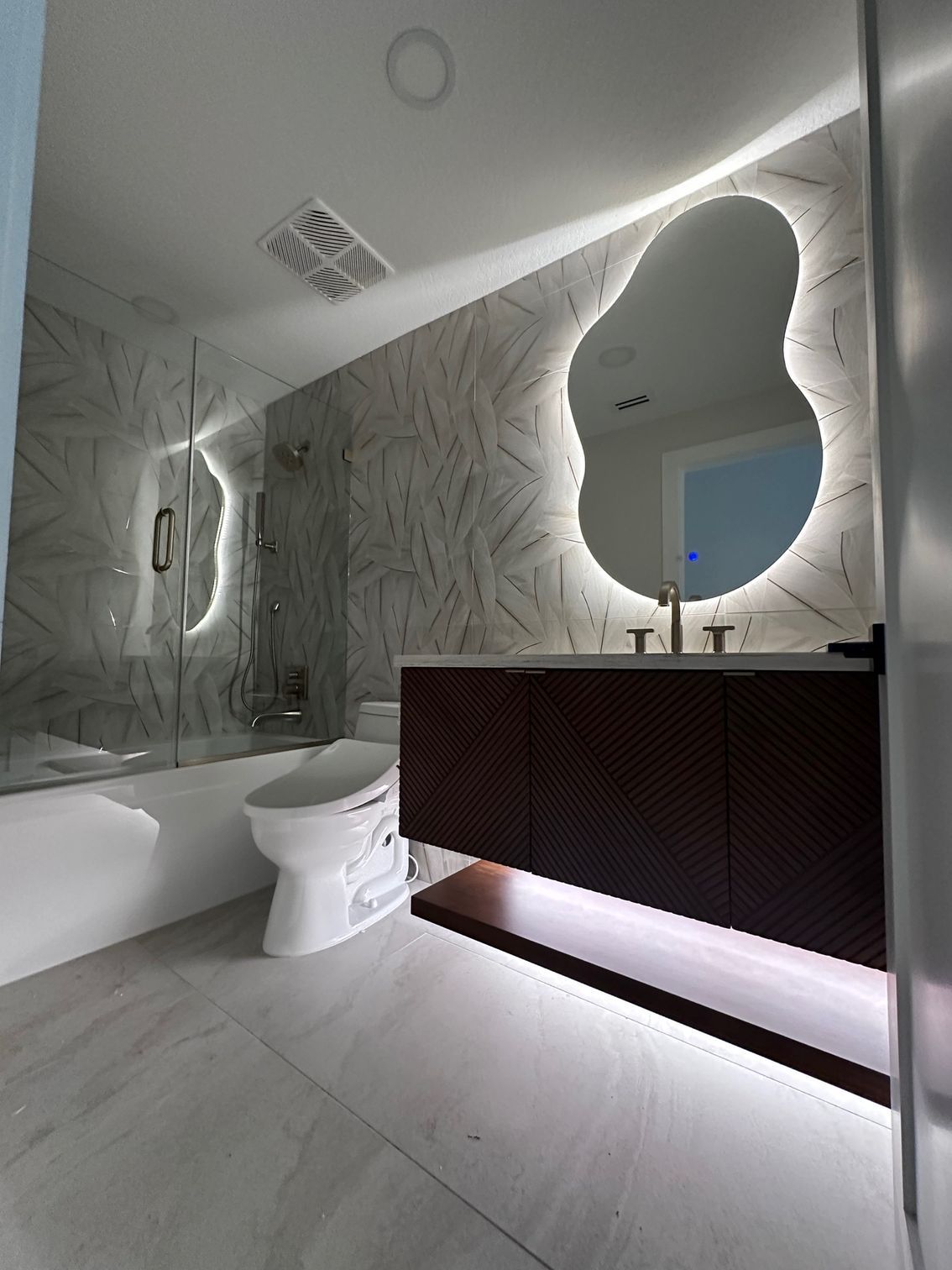 Modern bathroom with white tub, toilet, patterned wall, and illuminated dark vanity mirror.