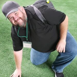 A man with a beard and a backpack is kneeling on the grass.