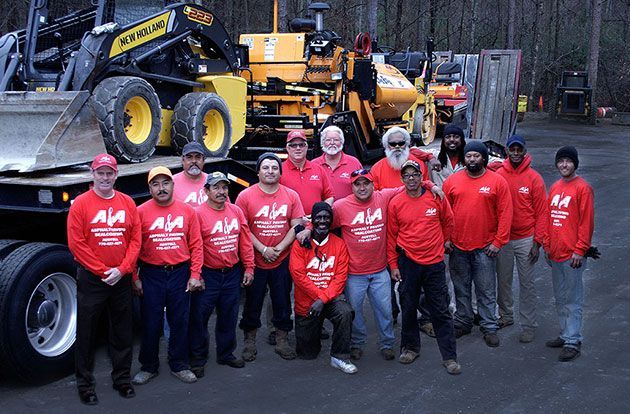 Group of men in red shirts with a logo pose with heavy machinery on a flatbed truck. They're outside on a cloudy day.