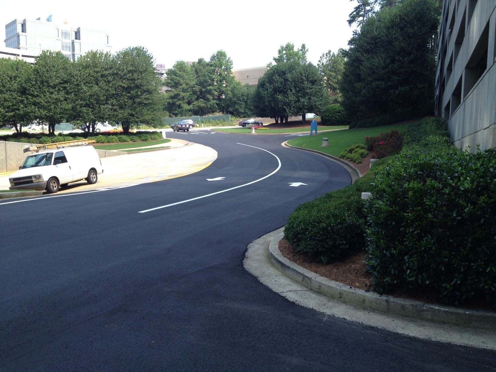 A paved road curves around a parking structure, marked with white lines. A white van is parked on the side. Green bushes and trees surround the area.