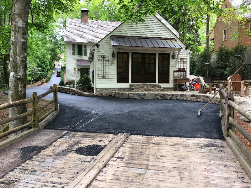 Asphalt paving on a wooden bridge leading to a white house with a dark-roofed porch. A split-rail fence and trees surround the scene.