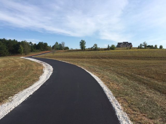 A paved road curves through a field with dry grass, leading towards a house on a hill under a blue sky.