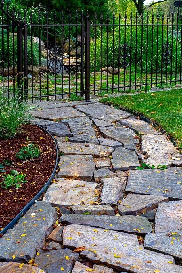 Flagstone path leading to a wrought iron gate and fence, flanked by a garden bed and lawn.