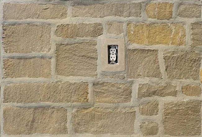 Stone wall with an electrical outlet centered. Beige and tan stones with gray mortar.
