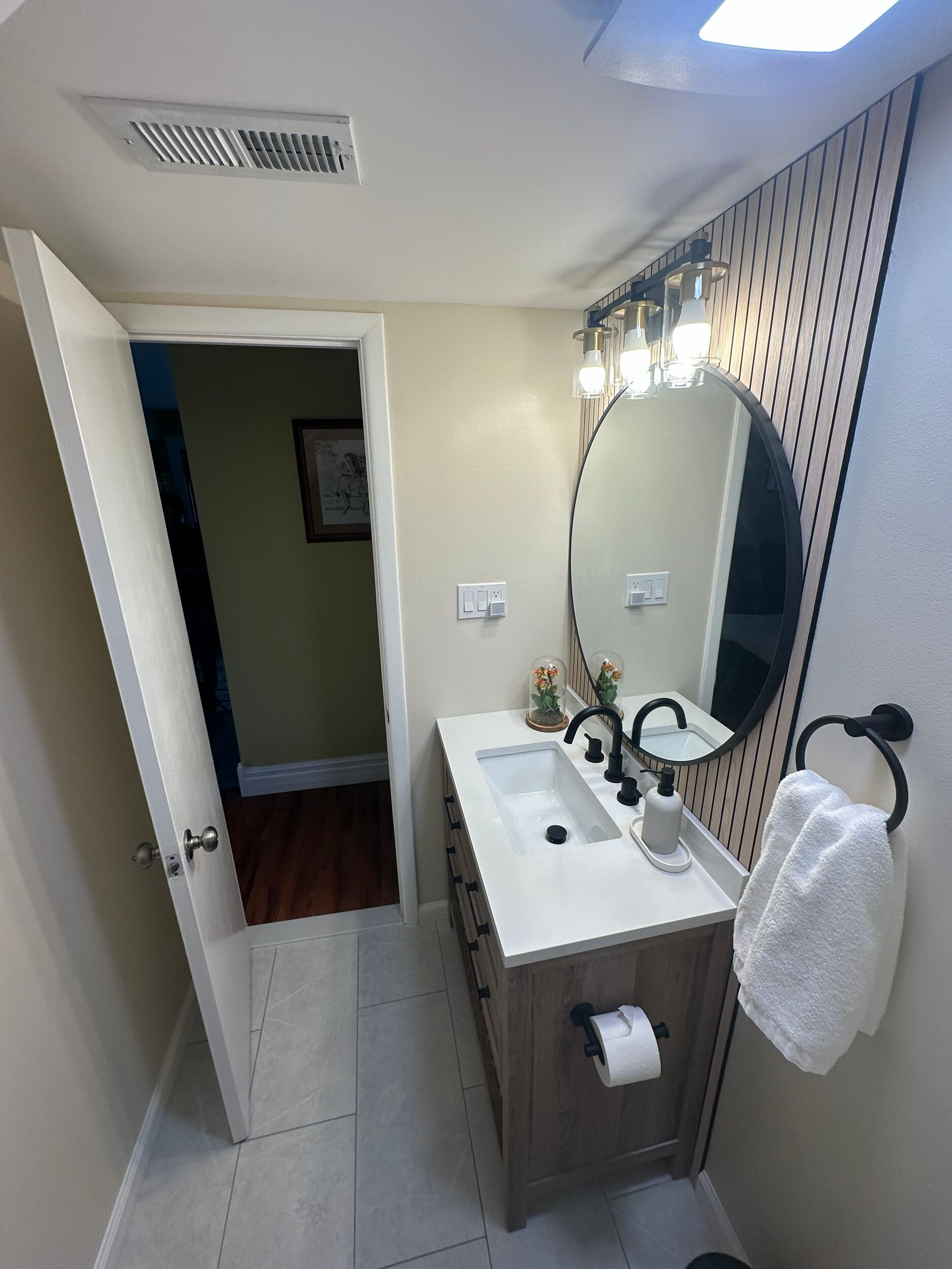 A modern bathroom vanity with a circular mirror, light fixture, and a white towel hanging on a black ring.