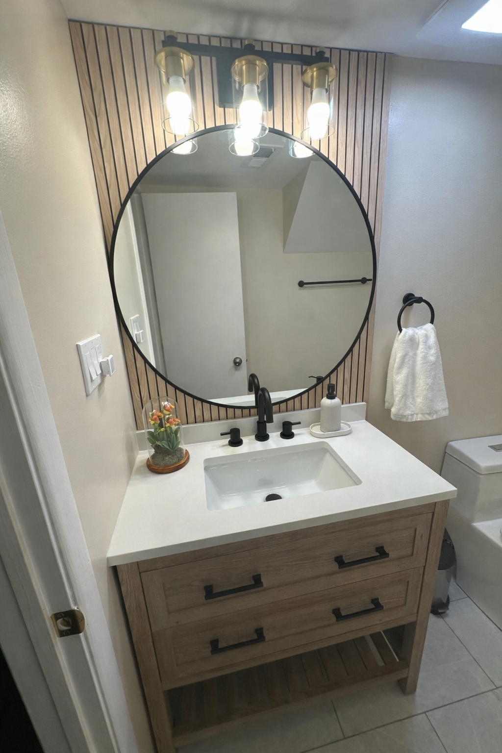 A modern bathroom vanity with a white countertop, round black-framed mirror, and wood-patterned accent wall.