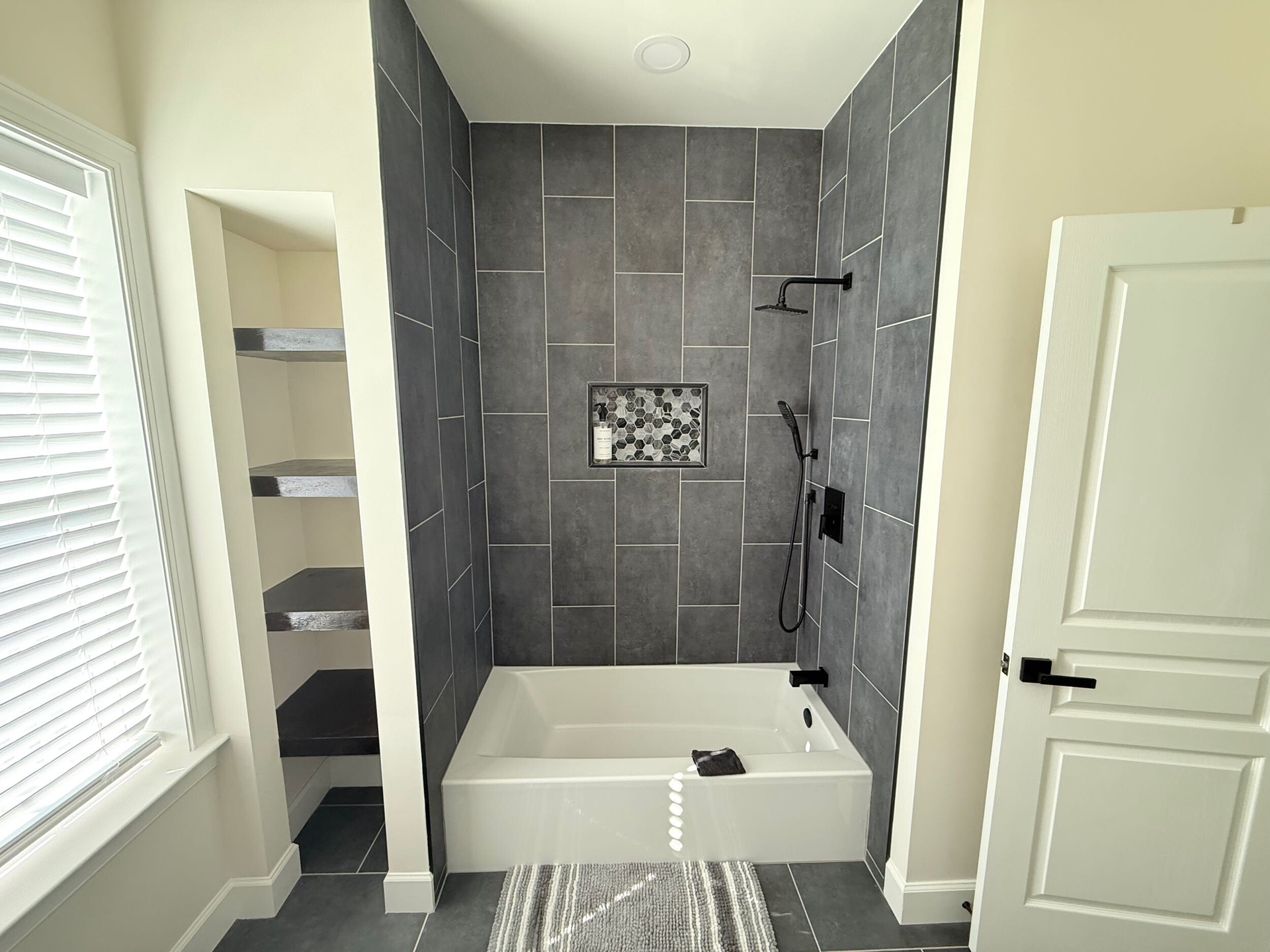 A modern bathroom featuring a white soaking tub set against a dark gray tiled wall, with open shelving beside it.